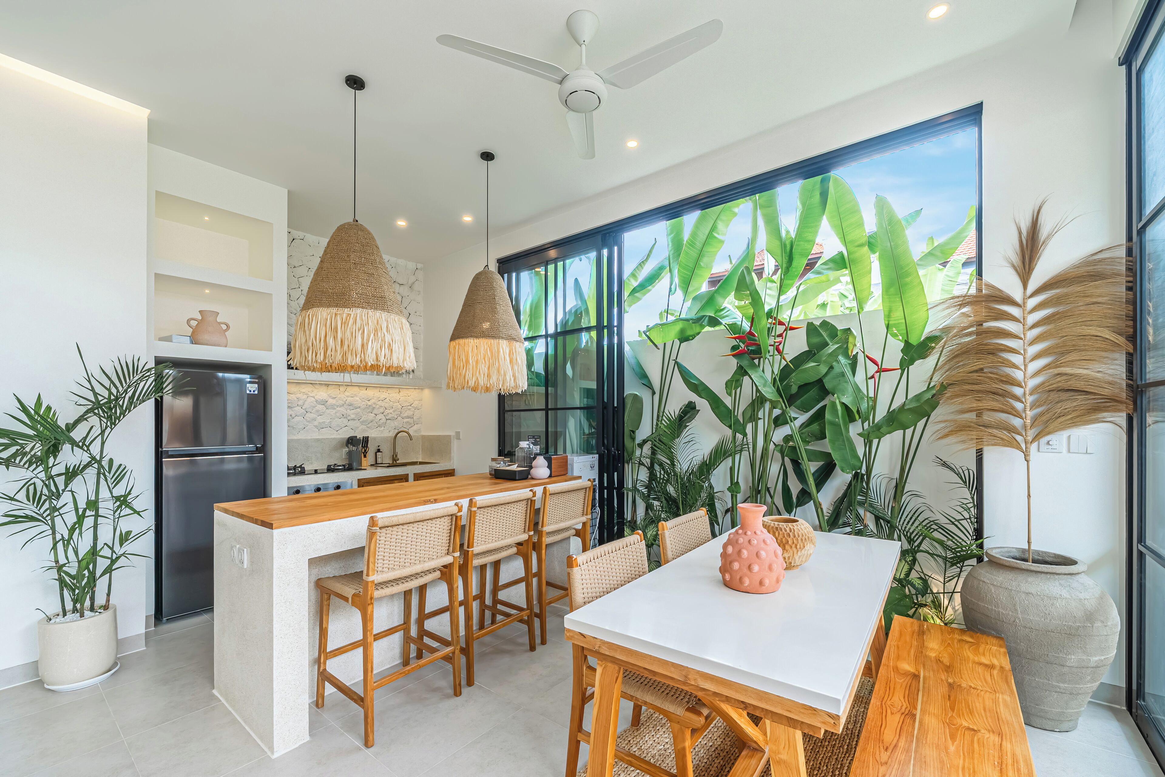A modern kitchen with a central island, wooden chairs, a white dining table, and large windows with plant views.