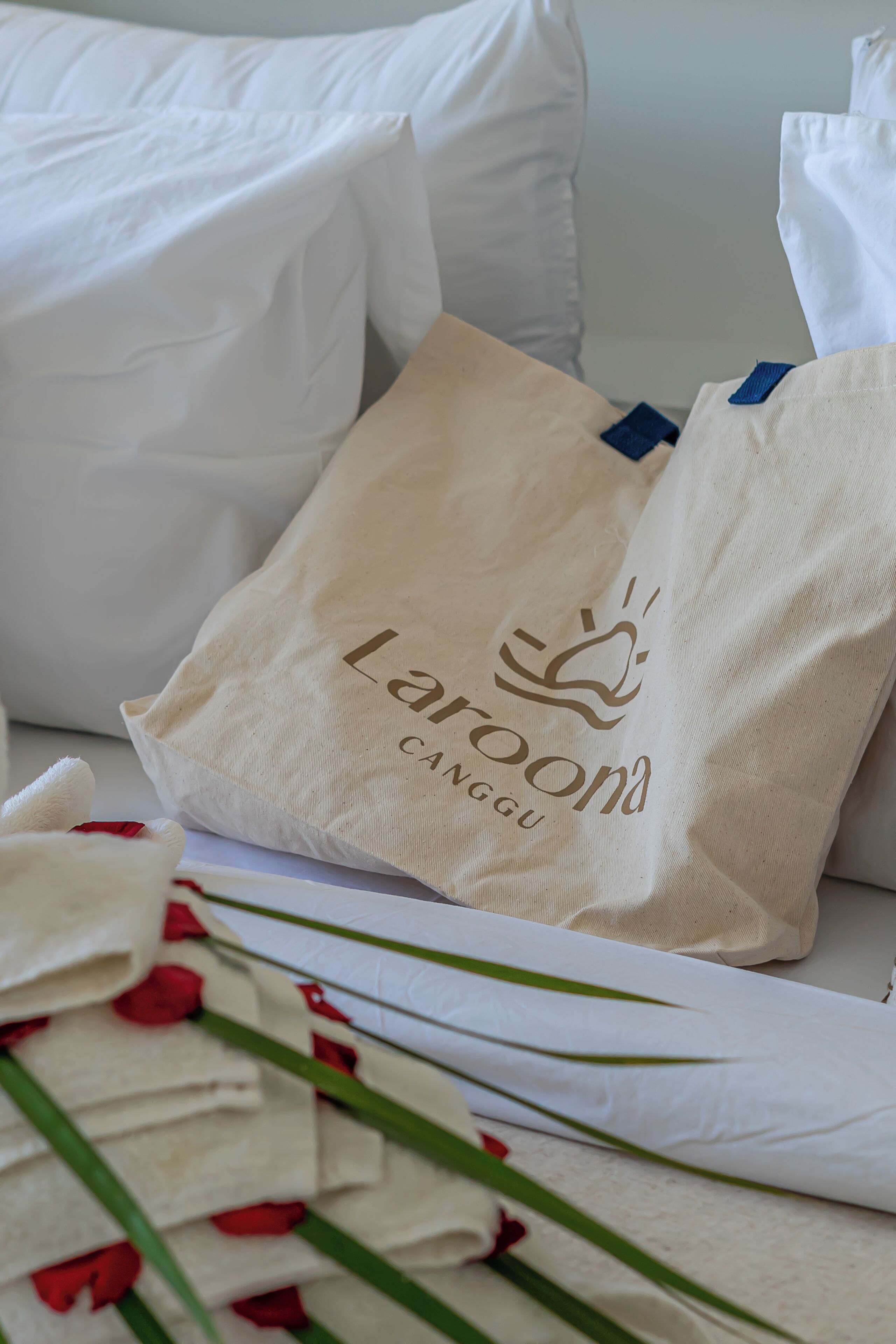 Eine beige Tragetasche mit dem Schriftzug 'Laraona' und einem Logo, auf einem Bett mit weißen Kissen und einer weißen Bettdecke platziert.