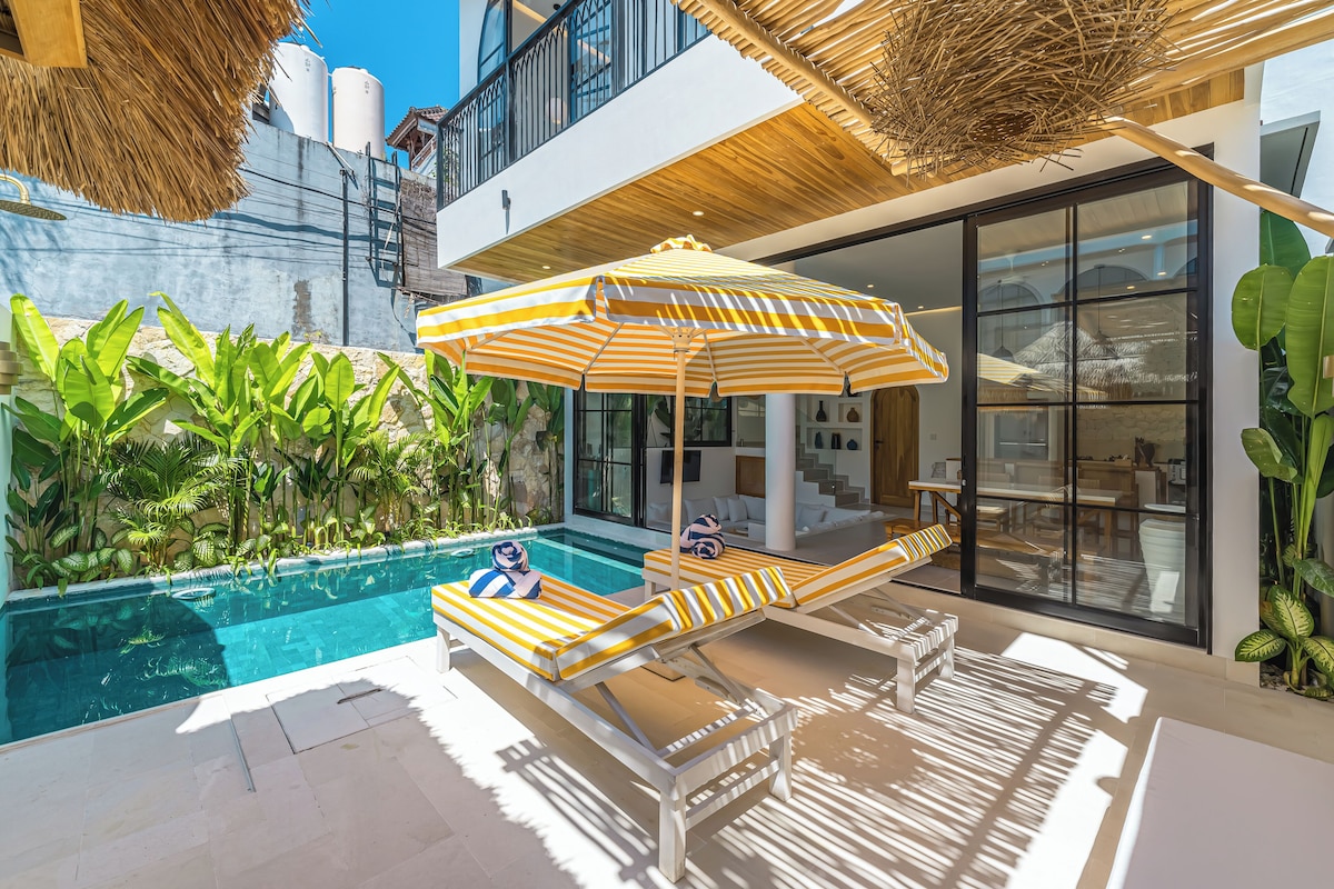 Un área junto a la piscina con sillones de descanso, una sombrilla y un edificio moderno con amplios ventanales y un balcón.
