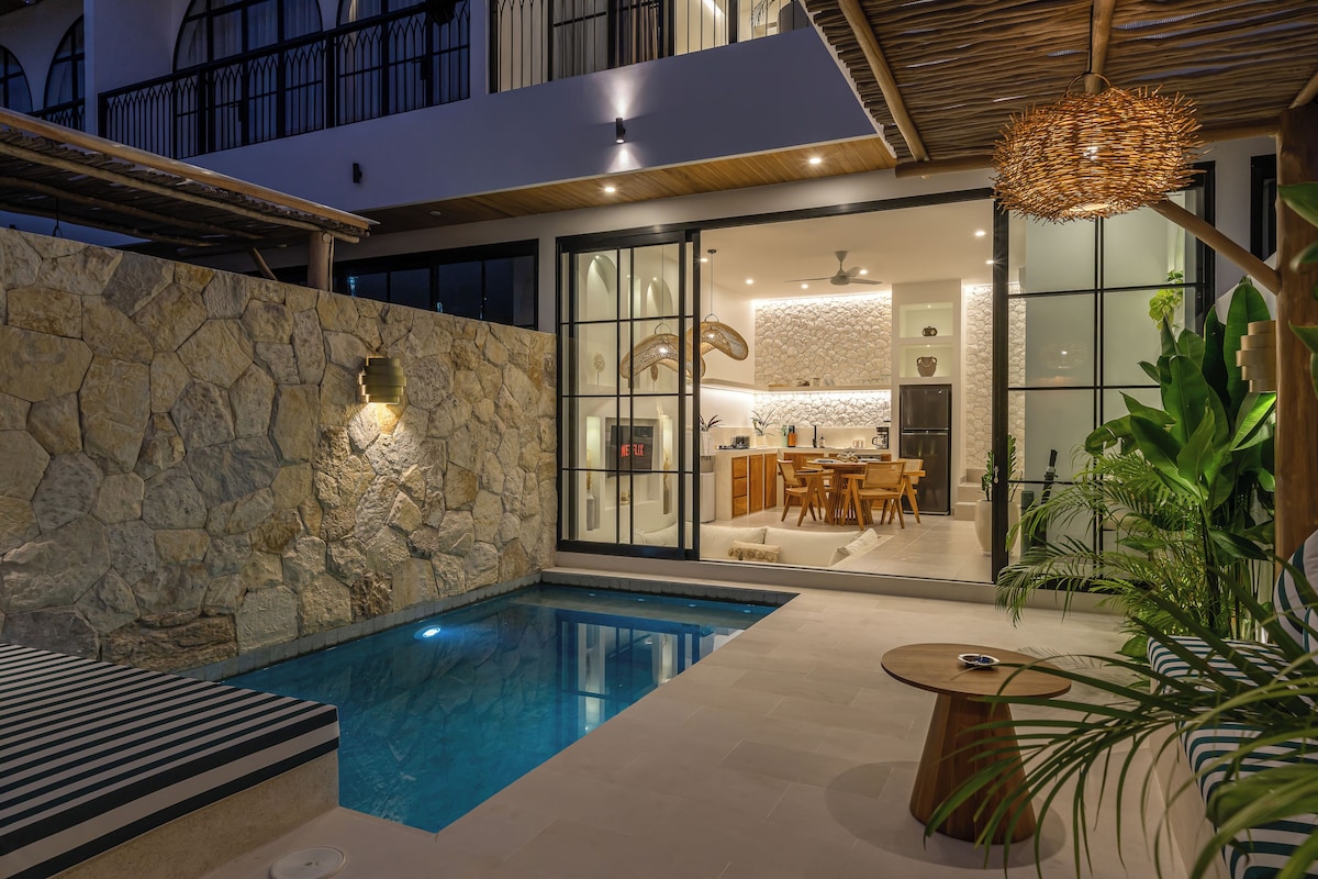 Un área de piscina moderna con una pared de piedra, una zona de comedor con cocina y un colgante luminoso.