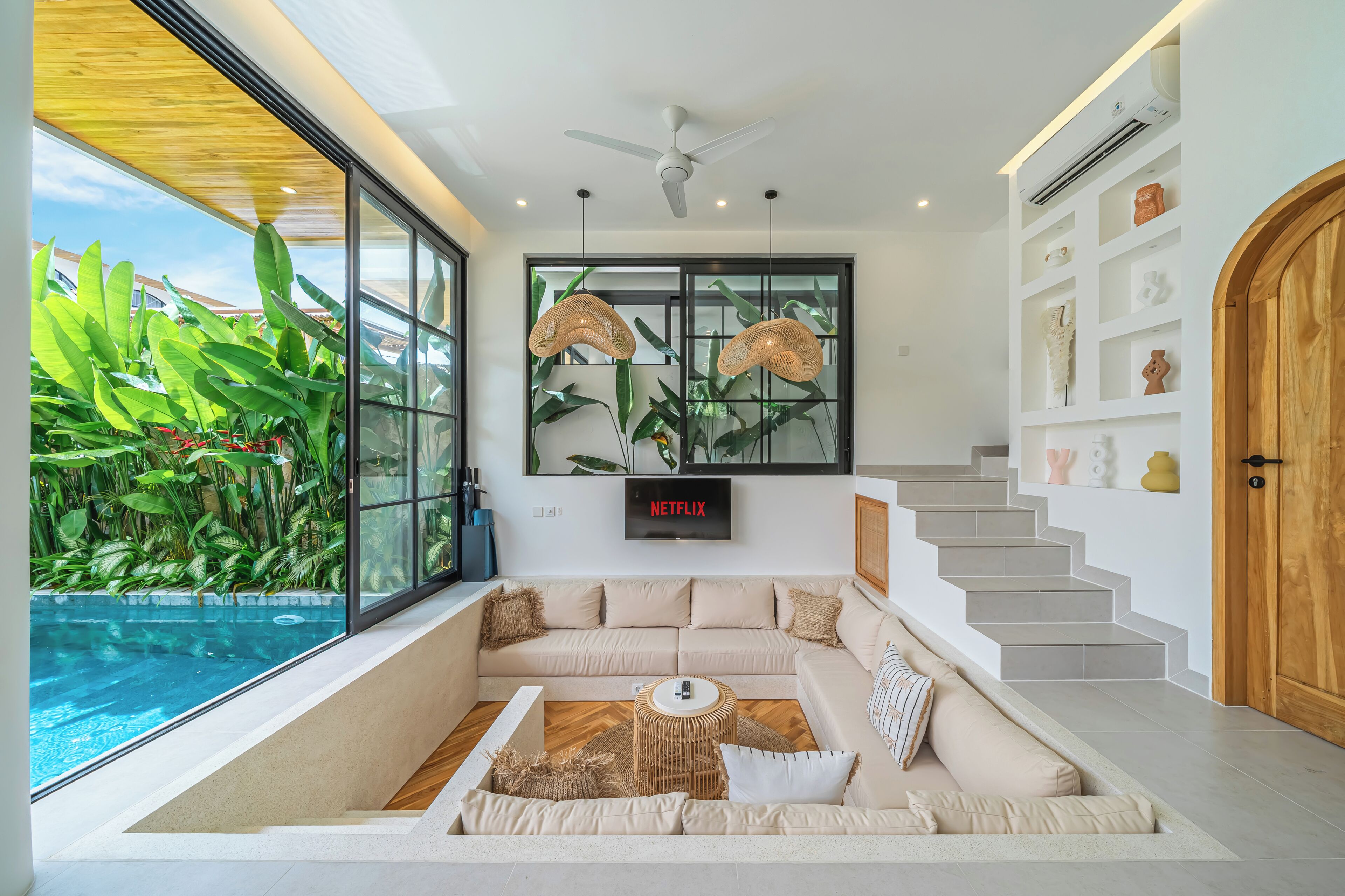 A modern living room with a large window, a ceiling fan, a flat-screen TV, a sofa, and a staircase.