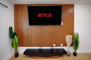 A modern living room with a wooden panel wall, a flat-screen TV mounted above a dark wooden coffee table, and two potted plants on either side.