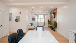 A modern living room with a white dining table, black chairs, a sofa, and a TV mounted on the wall.