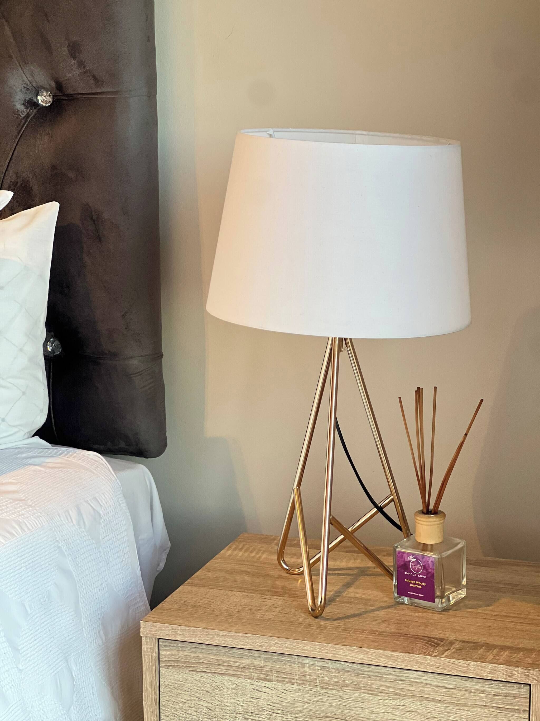 Ein Bett mit weißer Bettwäsche, ein dunkelbraunes Kopfteil, ein Nachttisch mit einer weißen Lampe und eine kleine Flasche Raumspray.