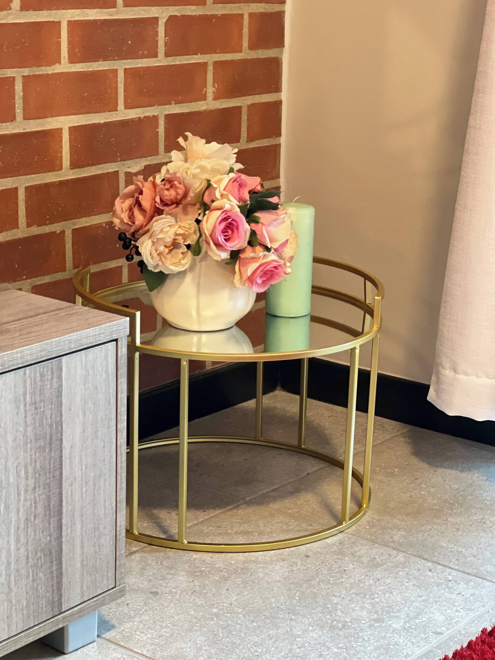 A round gold-framed side table with a white vase containing pink and peach flowers, positioned next to a brick wall and a white curtain.