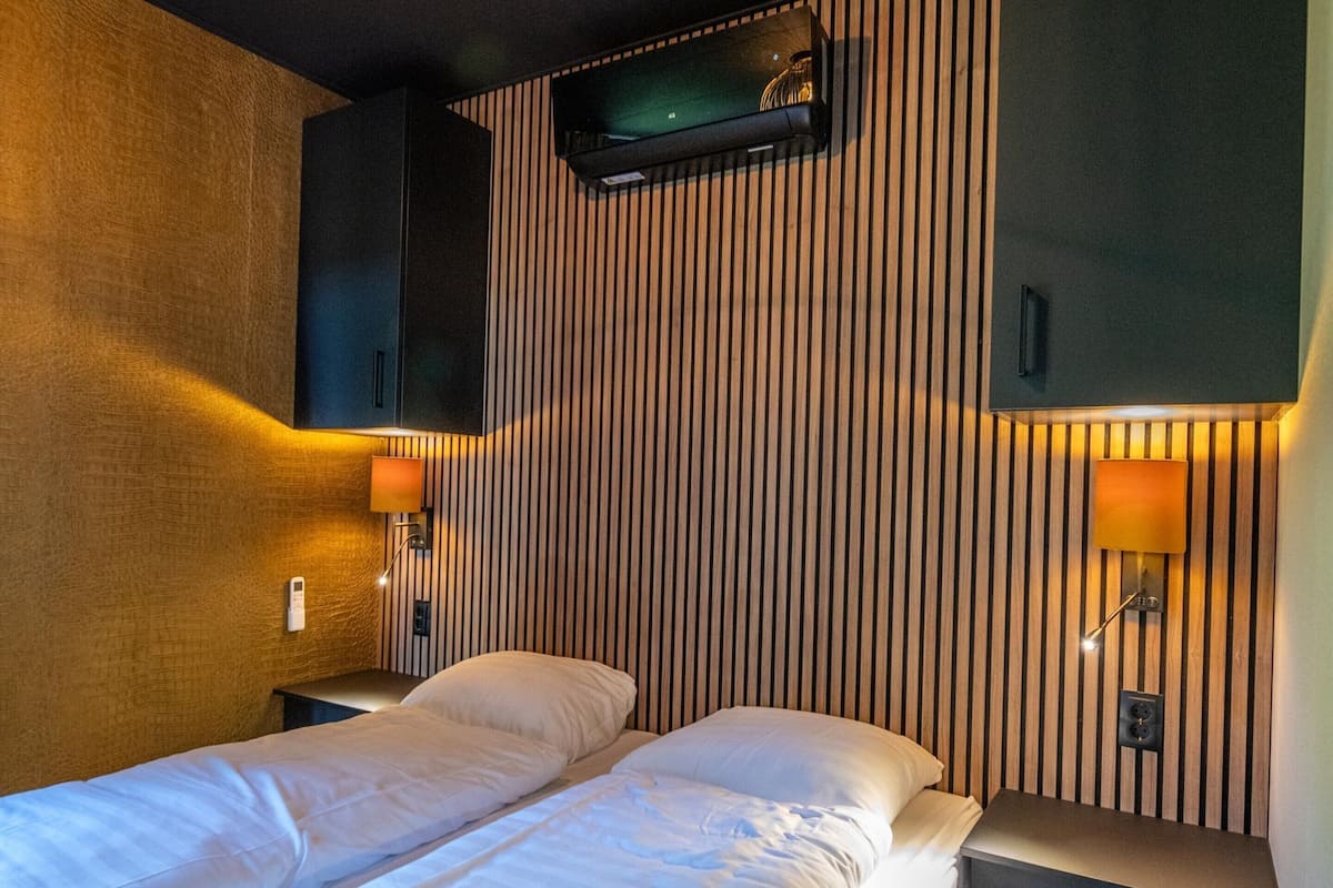 A hotel room with two single beds, a TV mounted on the wall, and two wall-mounted lamps.