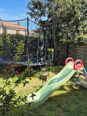 Ein Trampolin, eine Rutsche und ein eingezäunter Bereich mit Bäumen und einem Gebäude im Hintergrund.