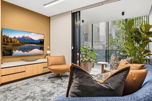 A modern living room with a large flat-screen TV, a sofa, a chair, and a balcony with plants.