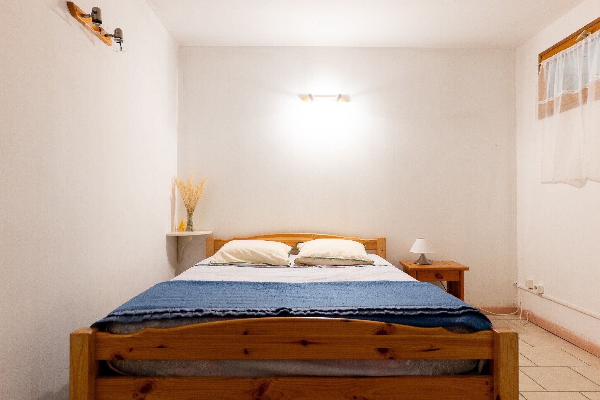 Une petite chambre à coucher, bien rangée, avec un lit en bois, une table de chevet avec une lampe et une étagère fixée au mur, ornée d’objets décoratifs.