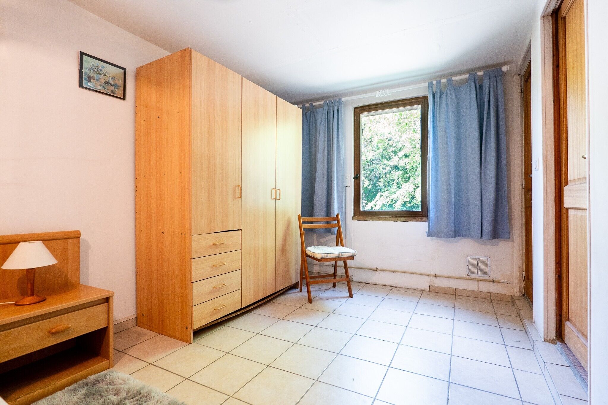 Ein Zimmer mit Holzschrank, einem Stuhl, einem Schreibtisch mit Lampe und einem Fenster mit Vorhängen.