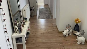 Käytävän lattia on parkettia, siellä on valkoinen penkki, jossa on koriste-esineitä, suuri peili ja ovi, joka johtaa toiseen huoneeseen.