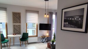 Ein modernes Wohnzimmer mit Fernseher, rundem Couchtisch, zwei grünen Sessel, einer Couch und einem gerahmten Bild an der Wand.