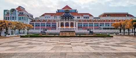 A large building with a central pavilion, a spacious paved area, and a row of trees.
