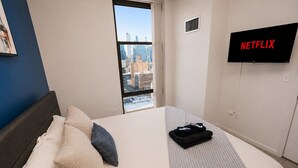 A hotel room with a bed, pillows, a blanket, a TV, and a city view.