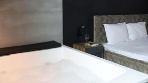 A modern bedroom with a large white bathtub, a tufted headboard, a bedside table with a phone and glass, and a neatly made bed. - Hotel Gloria KS (Pristina)