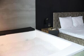 A modern bedroom with a large white bathtub, a tufted headboard, a bedside table with a phone and glass, and a neatly made bed. - Hotel Gloria KS (Pristina)