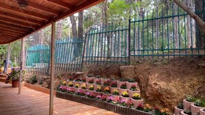 A covered wooden deck with a wooden railing, surrounded by a green fence and trees. - Hotel Gloria KS (Pristina)