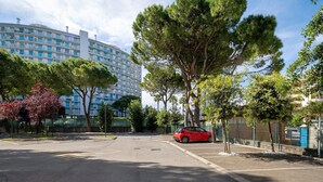 Un parking avec une voiture rouge, des arbres et, au fond, un grand immeuble à plusieurs étages.