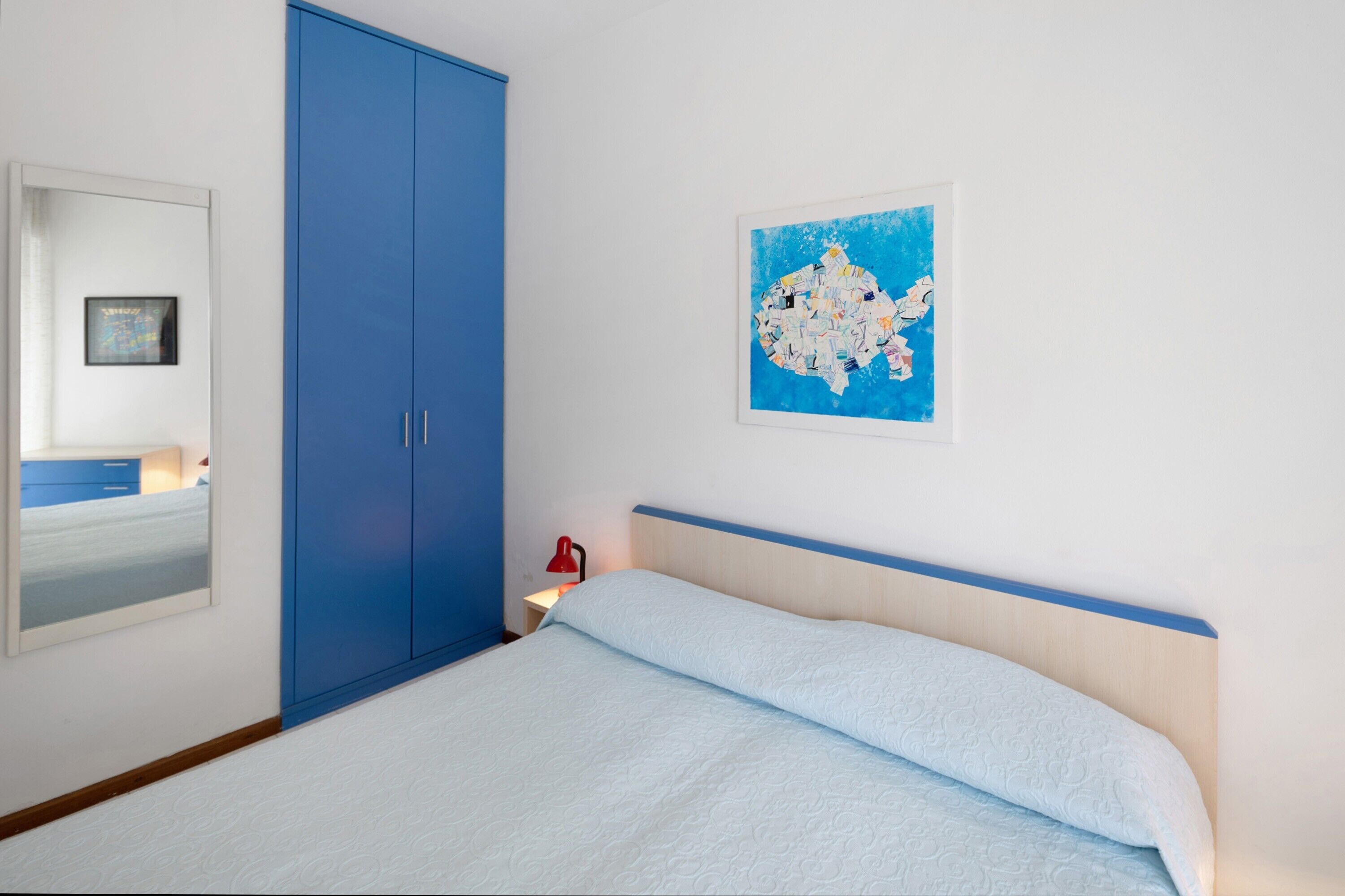 Une chambre à coucher avec un lit, une armoire bleue, une œuvre d’art accroch ée au mur et une table de chevet avec une lampe.