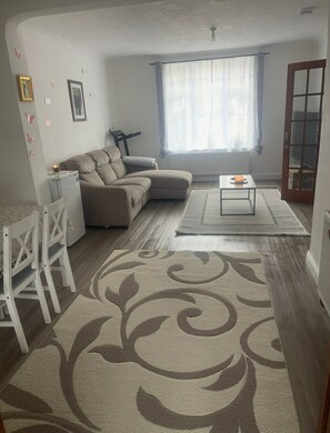 Een woonkamer met een bank, een salontafel, een eettafel met stoelen, een tapijt met patroon en een raam met gordijnen.