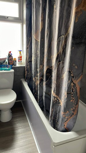 Un baño con un inodoro blanco, una bañera con azulejos negros y dorados desconchados y productos de limpieza en la encimera.