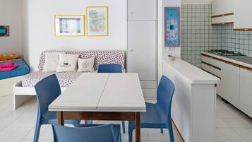 Une cuisine moderne avec des meubles blancs et un pare-feu en carrelage, un coin repas avec une table blanche et des chaises bleues, et un canapé agrémenté de coussins à motifs.