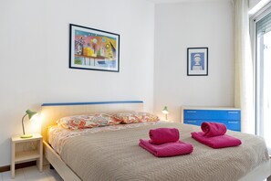 Une chambre d’hôtel avec un lit, une table de chevet avec une lampe, une commode bleue et deux œuvres d’art encadrées accrochées au mur.