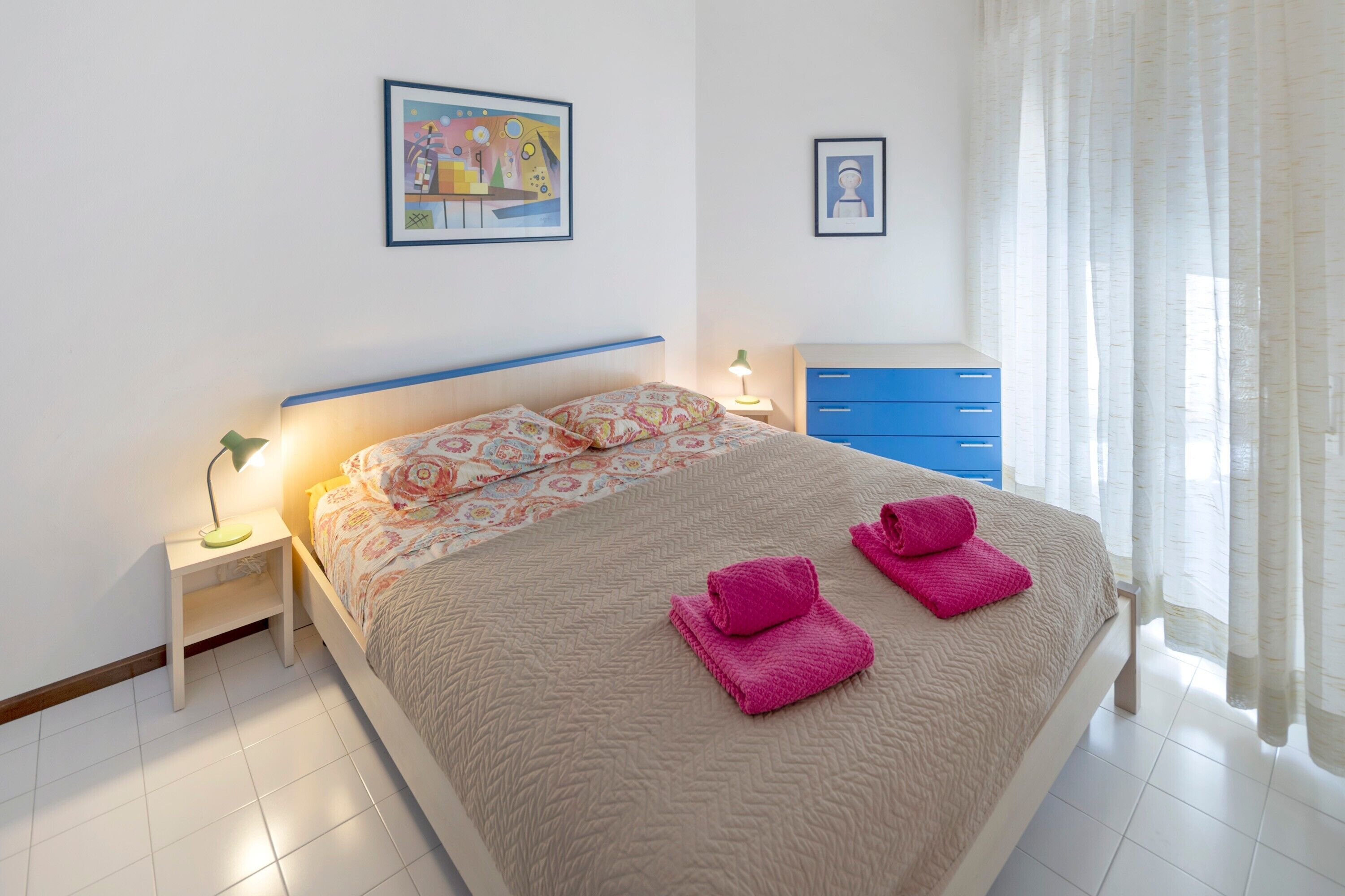 A neatly made bed with pink towels, a bedside table with a lamp, and a blue dresser in a bright room with white walls.