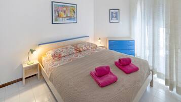A neatly made bed with pink towels, a bedside table with a lamp, and a blue dresser in a bright room with white walls.