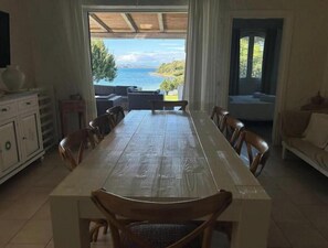 A dining area with a long table, chairs, and a view of the sea through a large window. - Beachfront villa with direct beach access - Costa Smeralda (Arzachena)