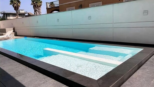 Ein rechteckiger Swimmingpool mit klarem, blauem Wasser, umgeben von einer gefliesten Terrasse.