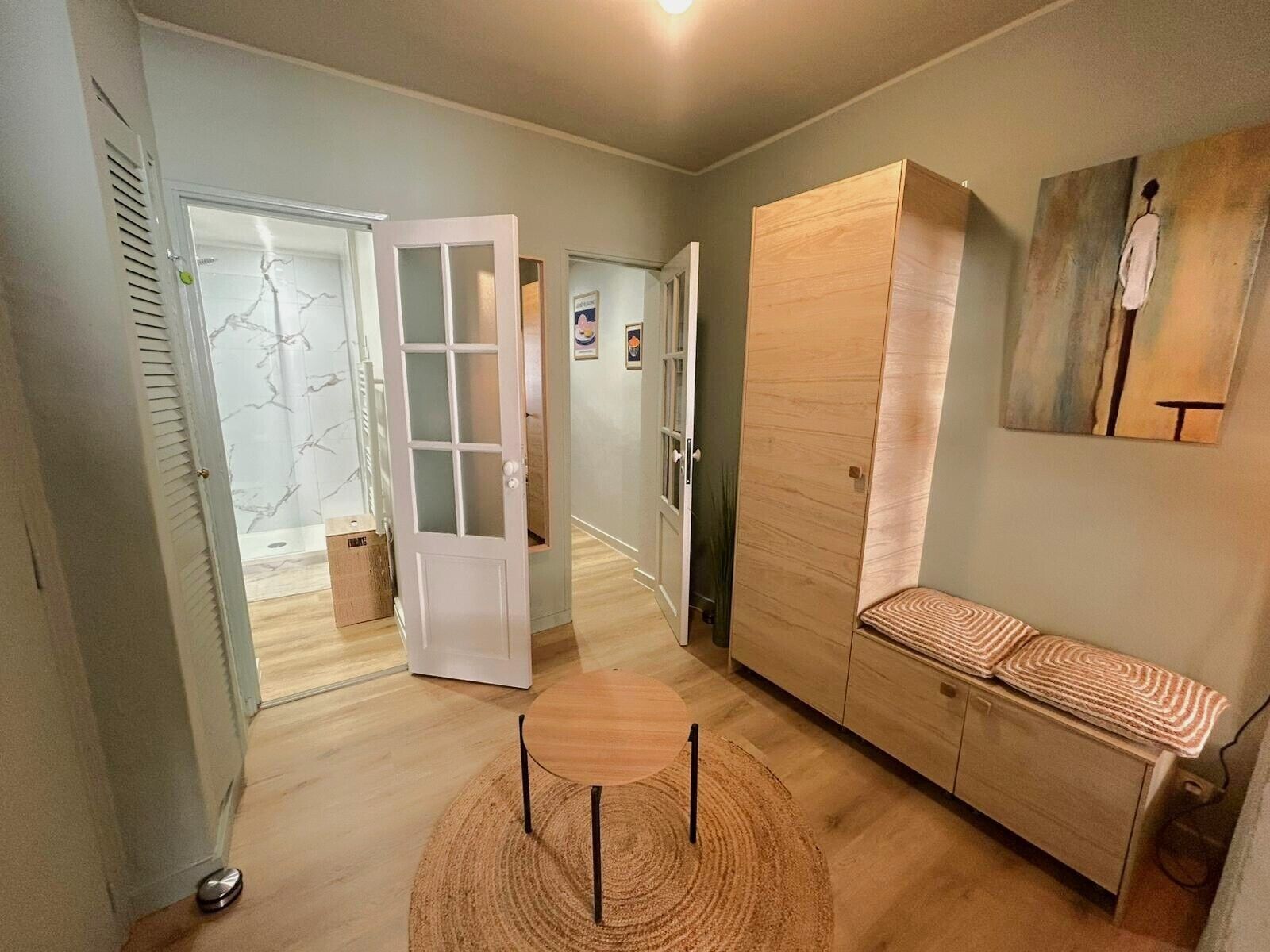 A hallway with a wooden floor, a white door, a wooden wardrobe, a bench, and a painting on the wall.