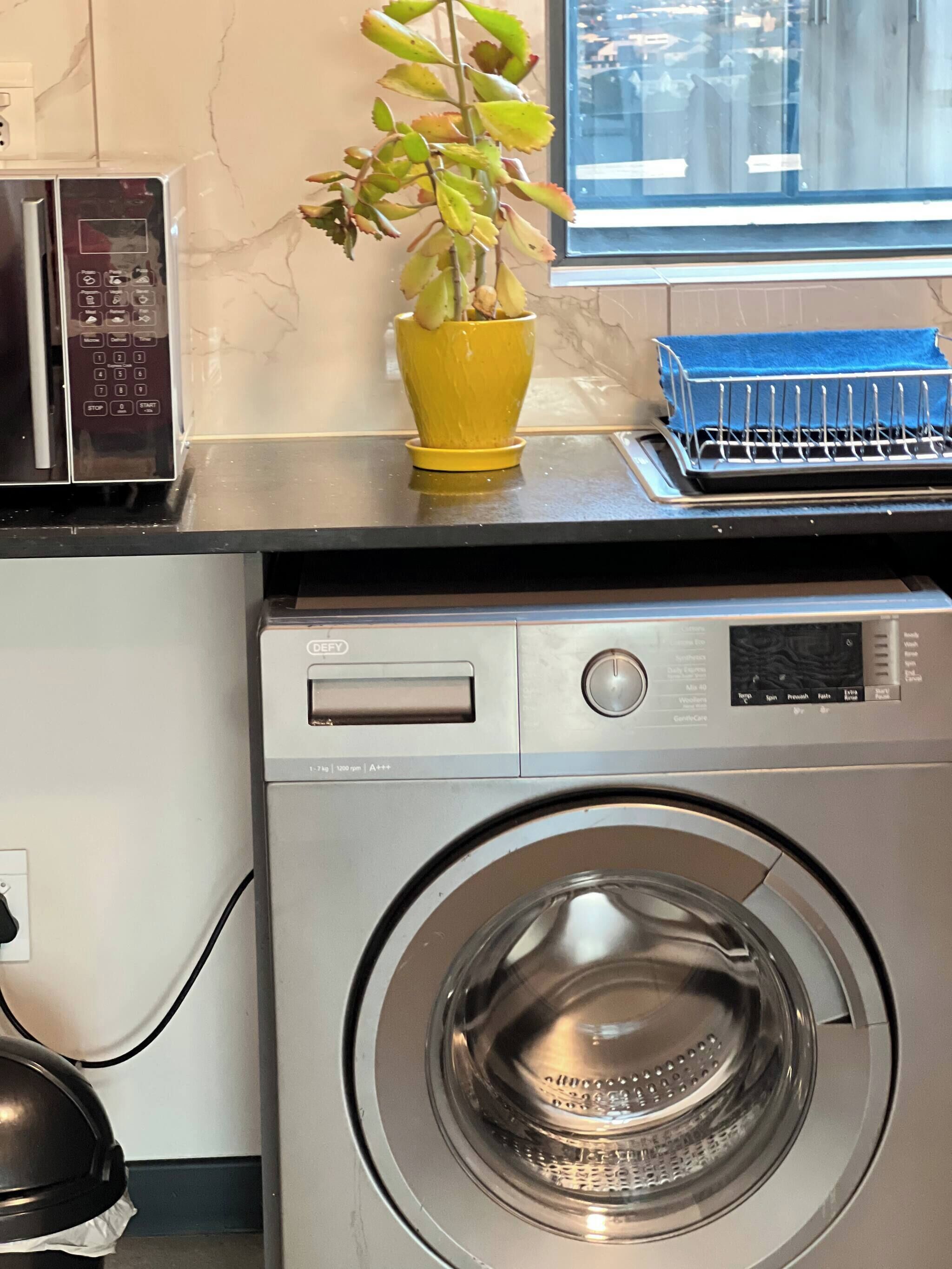Une cuisine compacte équipée d’un four à micro-ondes, d’un lave-linge et d’une plante en pot posée sur le plan de travail.
