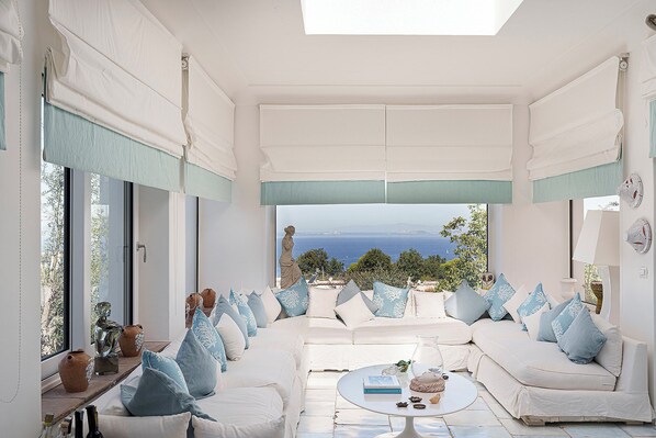 Ein sonnendurchflutetes Zimmer mit einem weißen Sofa, einem runden Tisch und Blick aufs Meer.
