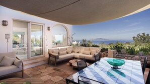 Um pátio externo com sofá, cadeiras, mesa e vista para o mar.