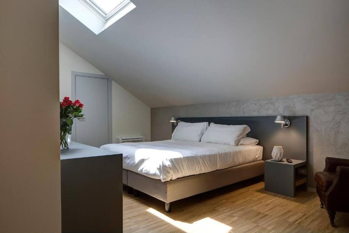 A bedroom with a skylight, a bed with white linens, a nightstand, a chair, and a vase with red flowers.