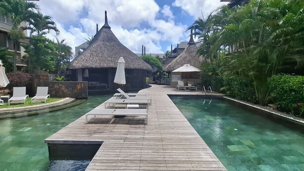 Un área de piscina en un complejo turístico con cubierta de madera, sillas de descanso y estructuras de techo de paja.