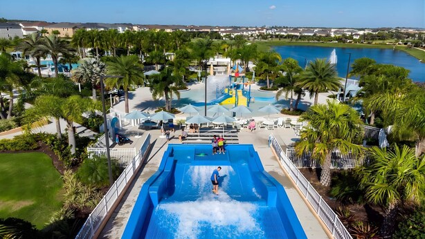 A water park with a large pool, palm trees, and a view of a lake and houses in the background. - Upstay Modern Home Near Disney With Pool (Kissimmee)