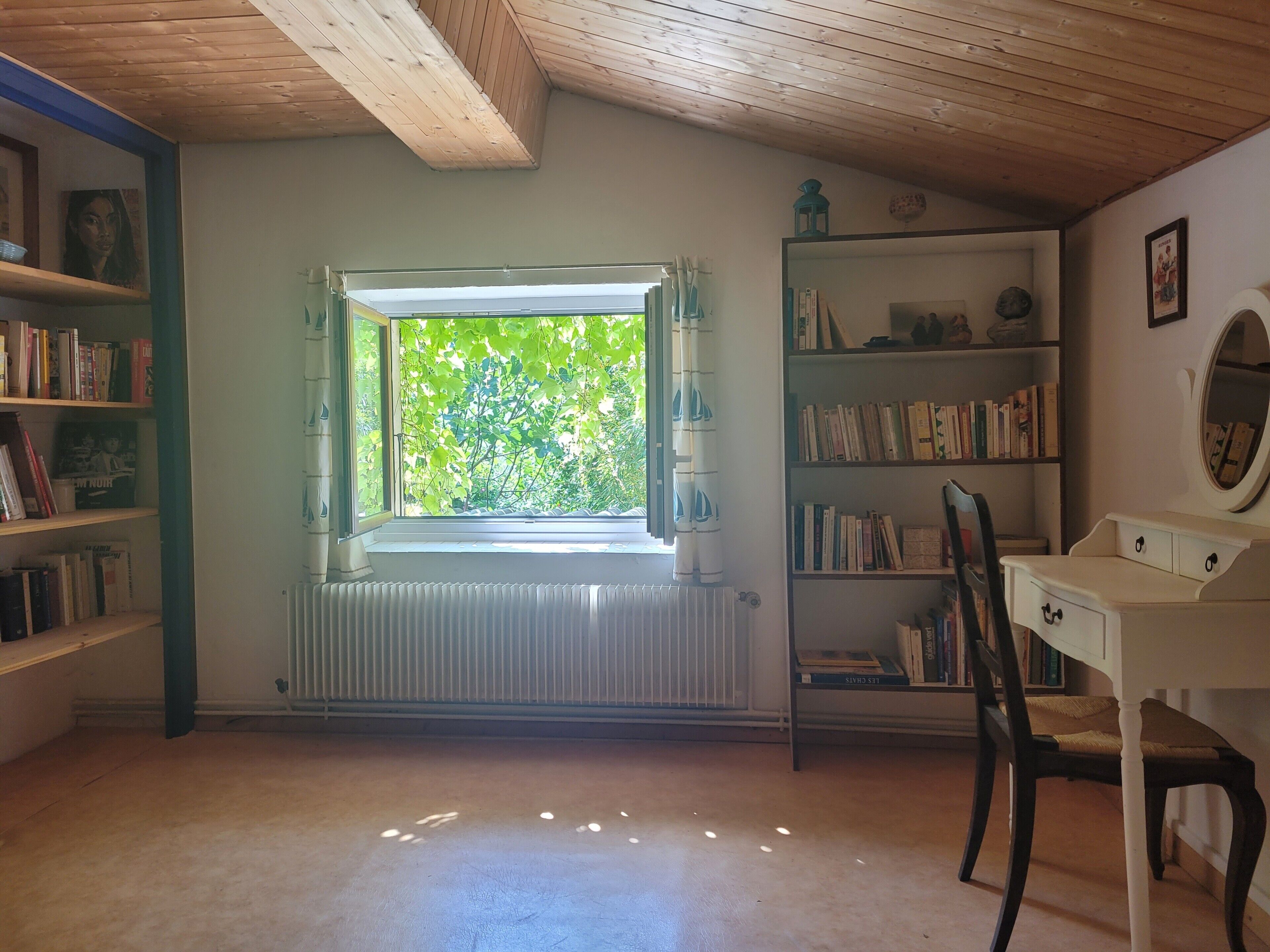 Ein Zimmer mit Holzbalkendecke, einem Fenster mit Blick auf Grünflächen, einem Heizkörper, einem Bücherregal und einem Schreibtisch mit Stuhl.