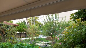 En udendørs patio med flisebelagt gulv, en stenvæg og udsigt til en have med forskellige planter og blomster.
