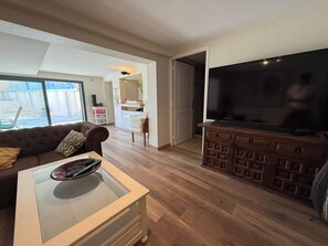 Ein Wohnzimmer mit einer Couch, einem Glastisch, einem Fernseher auf einem Holzkasten und Blick ins Freie.