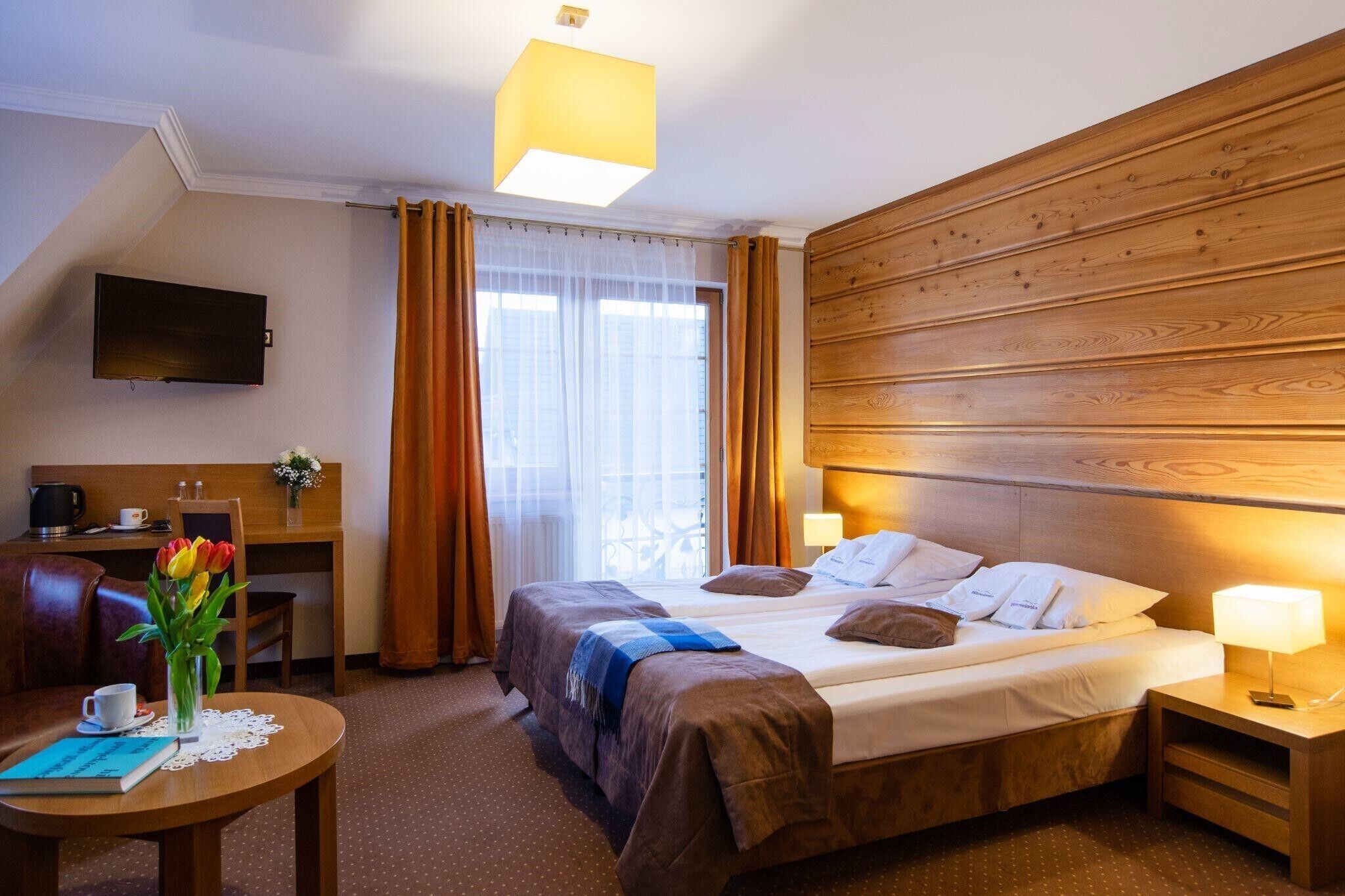 Una habitación de hotel con cama, mesitas de noche, escritorio, silla, televisor y ventana con cortinas.