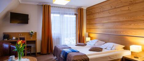 Una habitación de hotel con cama, mesitas de noche, escritorio, silla, televisor y ventana con cortinas.