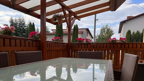 An outdoor patio with a glass table and wicker chairs, surrounded by wooden railings and potted flowers.
