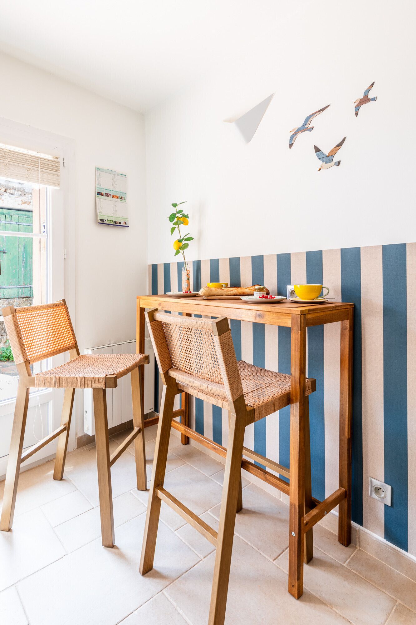 A room with a wooden table and two wicker chairs, a window with blinds, and a wall with a striped pattern and bird decals.