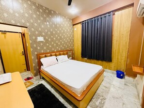 Habitación de hotel con cama, puerta de madera, ventana con cortinas, una mesita y un cubo azul.