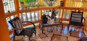 Una habitación iluminada por el sol con piso estampado, una silla de madera, un cojín a rayas, una mesita con una planta en maceta y una barandilla con vistas a la vegetación.