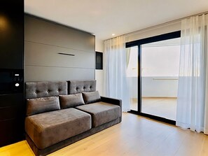 Ein modernes Wohnzimmer mit grauem Sofa, einem an der Wand befestigten Flachbildfernseher und einer Glasschiebetür, die auf einen Balkon führt.