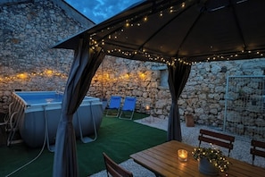 An outdoor patio with a stone wall, a hot tub, lounge chairs, and a wooden table with a candle. - Casa Valdebek With Pool (Pula)