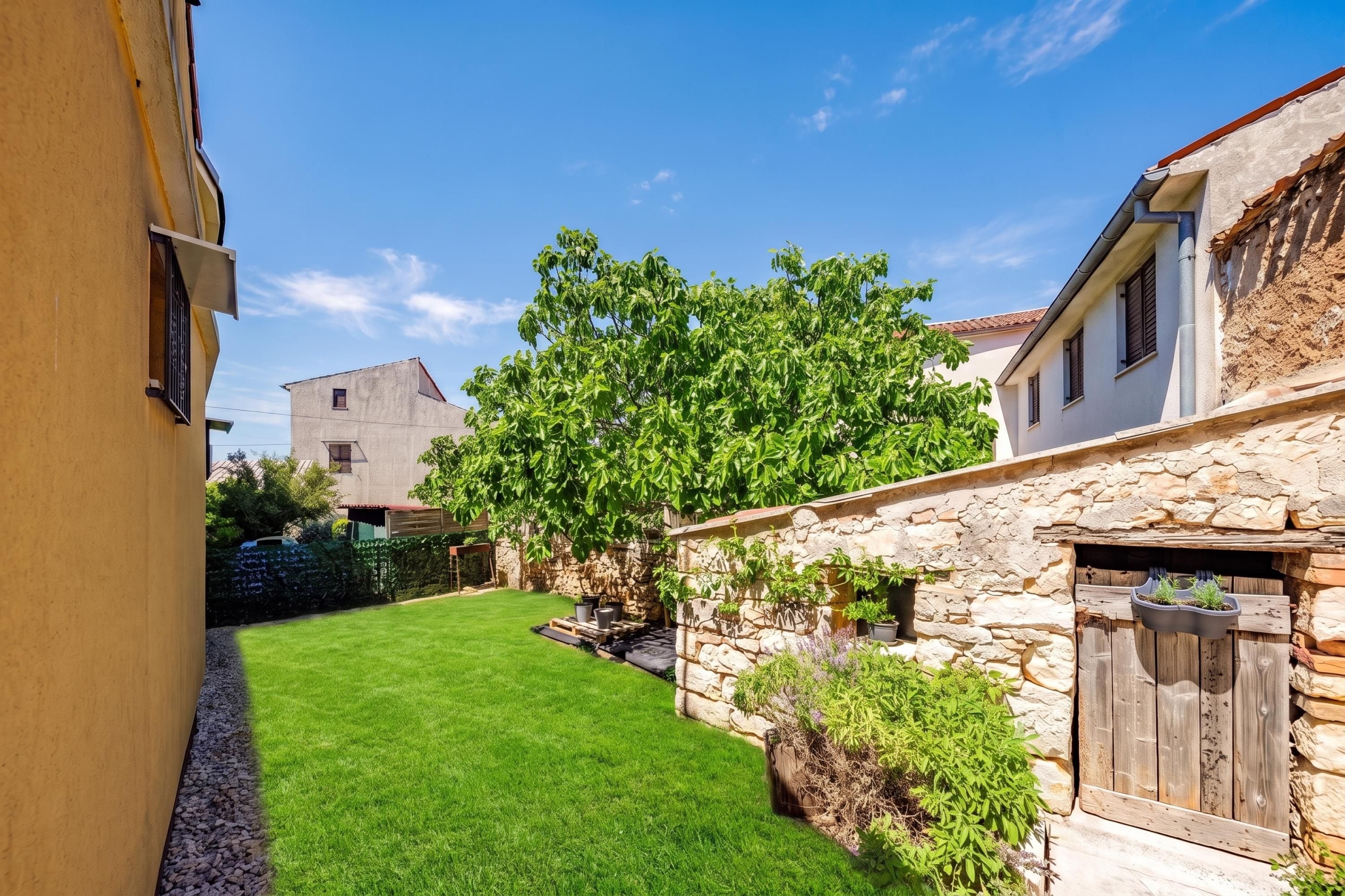 A narrow courtyard with a well-maintained lawn, stone walls, and a variety of plants.